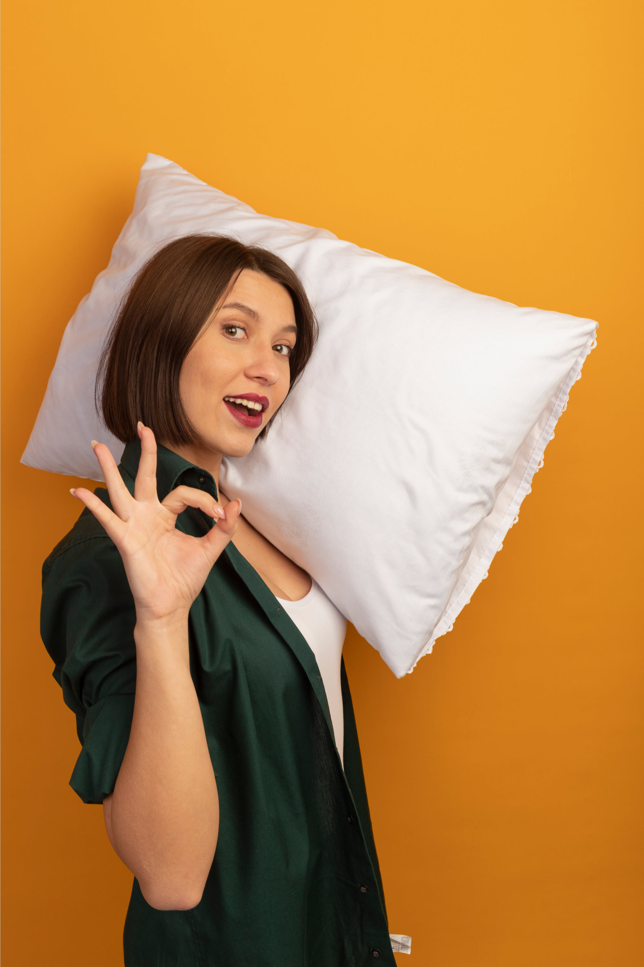 pleased pretty caucasian woman stands sideways holding pillow shoulder gesturing ok hand