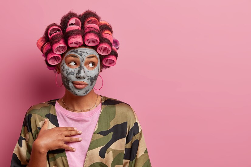 beautiful woman with hair curlers applies clay mask looks aside with thoughtful expression uses cosmetic product wears khaki dressing gown isolated pink wall blank space 273609 39527
